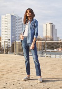 Jeansjack over een licht T-shirt, gecombineerd met donkere jeans en grijze instappers. Model staat op een houten terras met een stedelijke achtergrond.