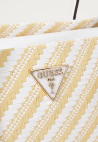 Woven torba w białym i żółtym kolorze, z trójkątną złotą blaszką z logo "GUESS USA". Teksturowana powierzchnia z wzorzystym wzorem.