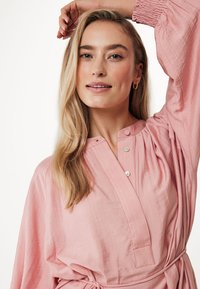 Blusa rosa chiaro con vestibilità rilassata, dotata di una patta con bottoni, maniche lunghe e un laccio in vita. Il tessuto testurizzato aggiunge interesse visivo.