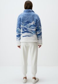 Giacca in shearling blu fuzzy con motivo a onda, interno in felpa bianca, colletto alto e vestibilità ampia. Abbinata a pantaloni jogger crema e sneakers.