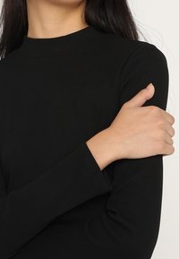 Musta ribbed pitkähihainen paita, jossa on korkea kaulus. Kangas näyttää sileältä ja se on muotoilultaan tiukka, esittäen lähellä vartaloa leikatut hihat ja vähäiset saumat.