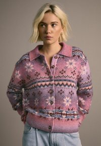 Kudotut neulecardigan vaaleanpunaisen ja purppuran sävyissä, joissa on geometrisia kuvioita. Varustettu nappikiinnityksellä, kauluksella ja ribbireunoilla.