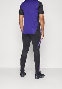 Maglietta sportiva viola e nera abbinata a leggings neri con dettagli viola e una chiusura lampo sul lato. Il materiale sembra elastico.