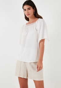 White blouse with a square neckline featuring lace detailing, short flutter sleeves, and a loose fit, paired with beige linen shorts.