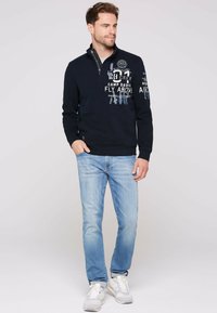 Sudadera de color azul marino con un cuello de media cremallera, que presenta texto gráfico en blanco. Combinada con vaqueros de color azul claro y zapatillas blancas.