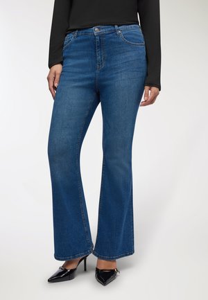 Donna che indossa jeans blue bootcut, un top nero a maniche lunghe e scarpe col tacco a punta nere su uno sfondo bianco.