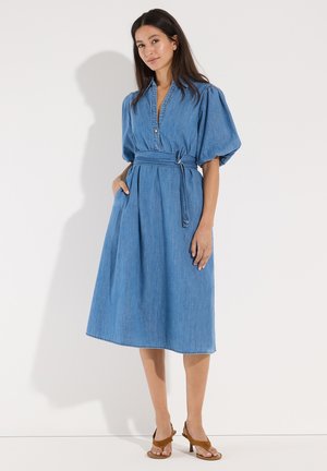 Frau trägt ein blaues Jeans-Midikleid mit Puffärmeln und Taillengürtel, steht mit einer Hand in der Tasche und braunen Sandalen mit Absatz.