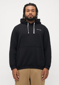 Bărbat cu barbă și dreadlocks îmbrăcat cu un hanorac negru cu șnururi albe și textul "CORE™ DEPT.", purtând pantaloni bej, stând în fața unui fundal alb.
