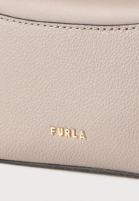 Close-up af beige tekstureret læder med gyldent Furla-logo og synlige syninger langs den øverste kant af materialet.