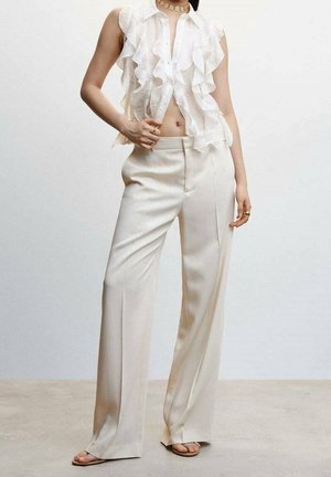 Woman wearing sleeveless white ruffled blouse, high-waisted cream wide-leg trousers, sandals, and a choker necklace, standing on a plain floor.