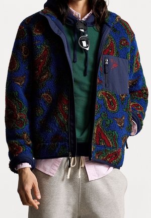 Person trägt eine blaue gemusterte Fleecejacke über einem grünen Kapuzenpullover und einem pinken Hemd, graue Sweatpants, mit Sonnenbrille, die an den Kapuzenbändern hängt.