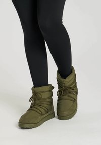 Botas verdes acolchadas con cordones en la parte delantera y cuello plegado, superficie texturizada y suela de goma. Llevadas con leggings negros.