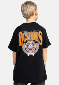 Černé tričko s velkým oranžovým textem "DCSHOES", kruhovým logem s vavřínem a nápisem "COAST TO COAST" nahoře, navržené pro uvolněný střih.