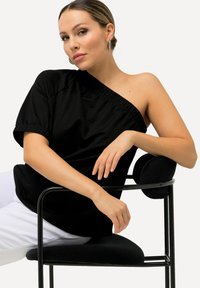 Sort off-shoulder top med samlet halsudskæring, afslappede ærmer og glat stof. Model siddende i en minimalistisk sort stol med cirkulær pude.