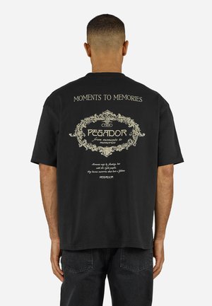 Mies seisoo selin, pukeutuneena mustaan T-paitaan, jossa on koristeellinen teksti "Moments to Memories" ja "Pegador" kermaan vivahtavalla värillä.