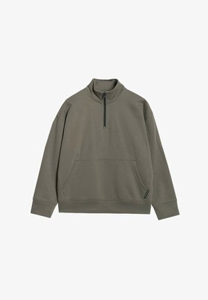 Enfant debout portant un sweat vert zippé oversize et un pantalon de jogging assorti avec des baskets sur fond uni.