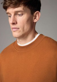 Sudadera marrón con un cuello redondo acanalado y tejido texturizado, superpuesta a una camisa blanca, exhibida sobre un fondo neutro.