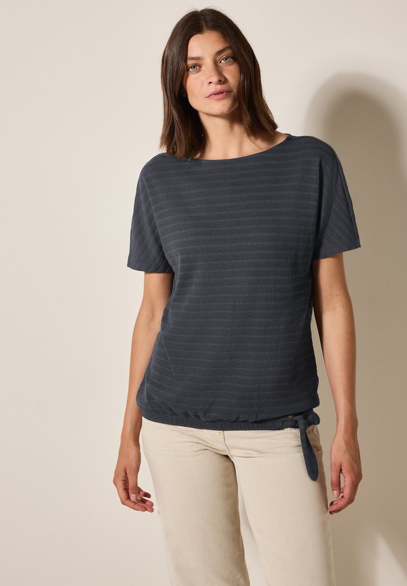 Haut gris foncé à manches courtes avec un motif texturé et rayé, doté d'un détail de ourlet noué. Associé à un pantalon léger à jambe droite.