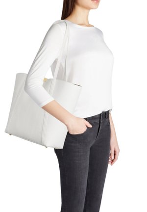Donna che indossa una maglia bianca a maniche lunghe e jeans neri, con una grande borsa bianca a tracolla, mano in tasca, su sfondo bianco.