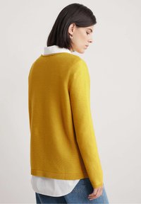 Gul ribbad tröja med långa ärmar, rundad halsringning och avslappnad passform, lager-på-lager ovanpå en vit skjorta med krage, kombinerad med denimjeans.