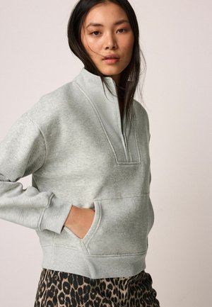 Grauer Pullover mit langen Ärmeln und halber Reißverschluss, geripptem Kragen und vorderer Kängurutasche. Getragen mit einer Hose im Leopardenmuster.