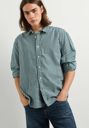 Junger Mann mit schulterlangem braunem Haar trägt ein grün-weiß kariertes Hemd mit hochgekrempelten Ärmeln und blaue Jeans, Hände in den Taschen.