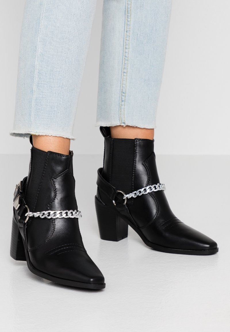Bottes cheville en cuir synthétique noir avec un bout pointu et un talon bloc. Présentent un accent de chaîne argentée autour de la cheville et des côtés élastiques.
