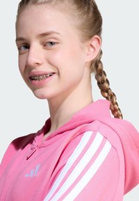 Sudadera rosa con cremallera y rayas blancas en los hombros. Presenta un corte relajado y una tela suave. El modelo tiene el cabello trenzado y lleva frenillos.