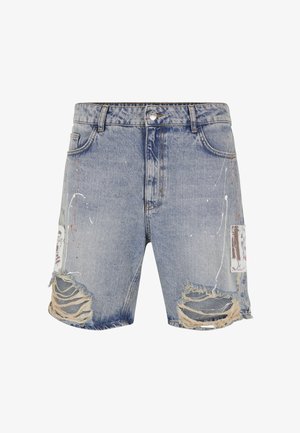 Homme portant un short en jean bleu clair déchiré avec des éclaboussures de peinture, des chaussettes blanches et des baskets blanches, debout sur un fond uni.