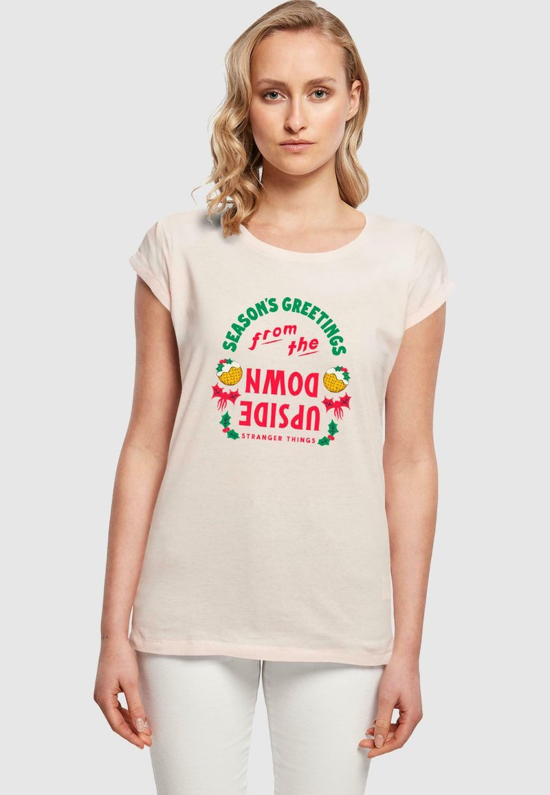 Rosa t-shirt med kort ärm som har en färgglad grafik med texten "Säsongens Hälsningar från Upside Down" tillsammans med juligare inslag och "Stranger Things"-text.