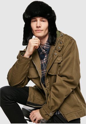 Giacca verde oliva con numerose tasche, chiusura con zip, e un cappello trapper in pelliccia nera. Il modello indossa anche una camicia a quadri e pantaloni neri.