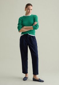 Maglione verde a trecce con maniche lunghe indossato sopra una camicia bianca, abbinato a pantaloni blu scuro e scarpe slip-on a fantasia.