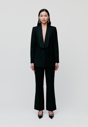 Black tailored suit featuring a deep V-neckline and wide-legged trousers. Glossy black pointed-toe heels and chandelier earrings accent the look.