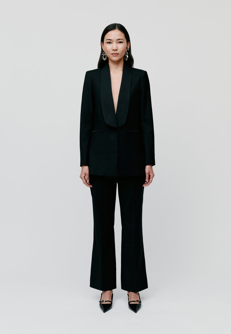 Black tailored suit featuring a deep V-neckline and wide-legged trousers. Glossy black pointed-toe heels and chandelier earrings accent the look.