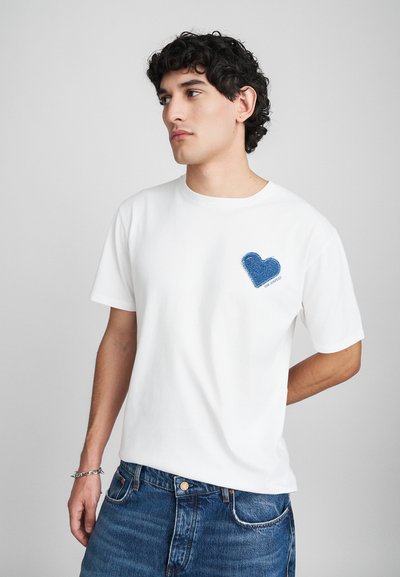 Weißes Baumwoll-T-Shirt mit einem blauen Denim-Herz-Patch auf der Brust. Das Design umfasst einen Rundhalsausschnitt und kurze Ärmel.