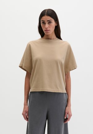 Woman with long dark hair wearing a loose beige short-sleeve top and gray wide-leg pants standing against a plain light background.