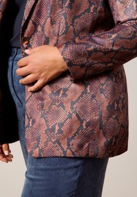 Blazer imprimé serpent marron et noir avec une texture lisse, doté d'un seul bouton doré. Associé à un pantalon en velours côtelé bleu foncé.