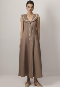Vrouw in een mouwloze lange bruine jurk met brede kraag en strik aan de voorkant, gecombineerd met beige slingback schoenen, staande tegen een effen achtergrond.