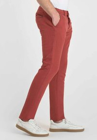 Man med slim-fit rustfärgade byxor och vita sneakers, stående med händerna i fickorna mot en enkel bakgrund.