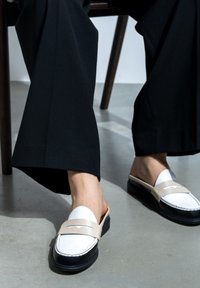 Beige und weiße Slipper mit schwarzer Sohle, mit einem dekorativen Riemen und einer Stichdetail. Getragen mit lockeren schwarzen Hosen.