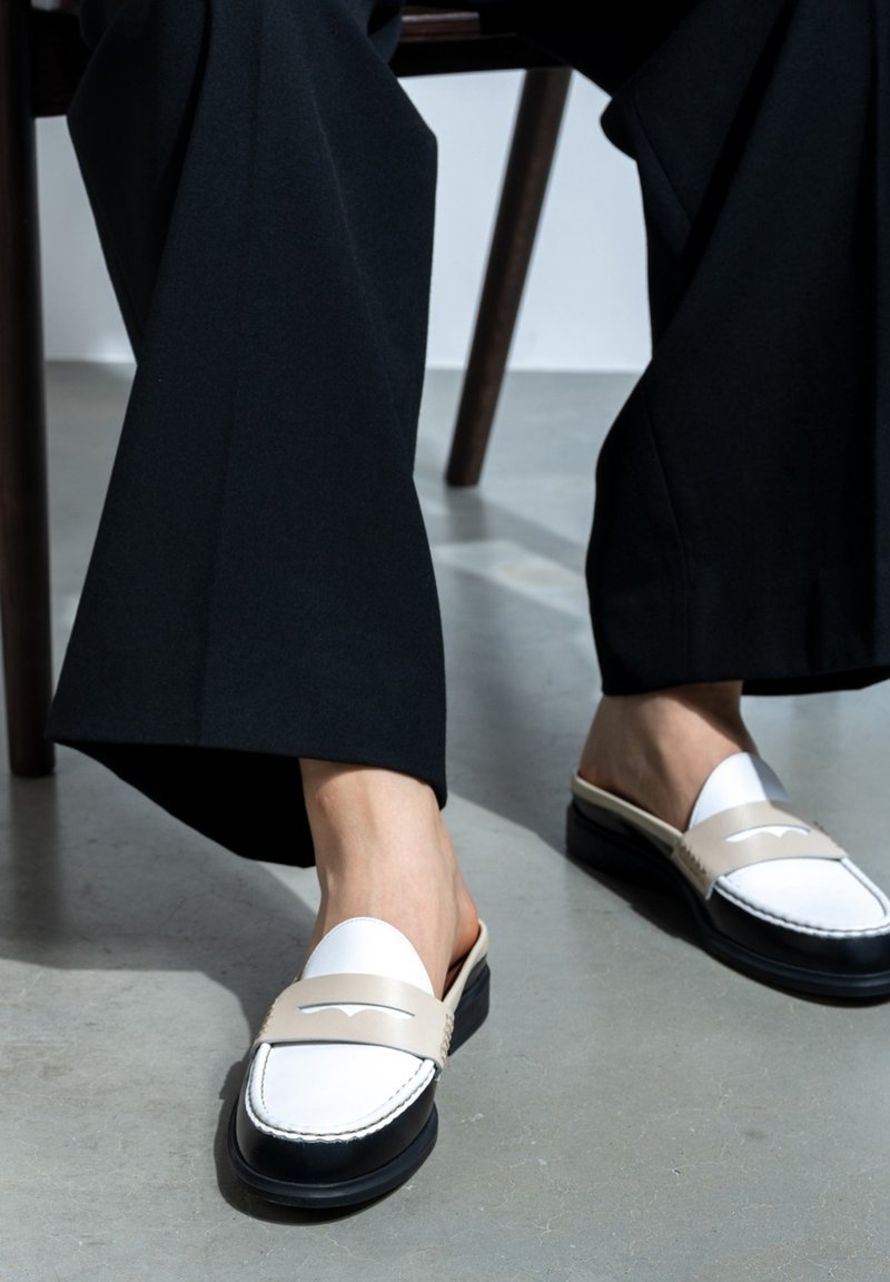 Beige en witte instaploafers met een zwarte zool, voorzien van een decoratieve band en stikseldetail. Gecombineerd met losse zwarte broeken.