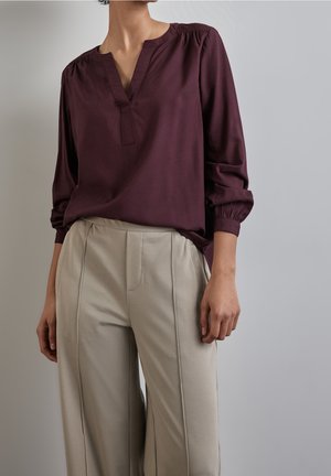 Osoba ubrana w długą bluzkę z głębokim dekoltem w kształcie litery V w kolorze głębokiego fioletu oraz beżowe, wysokie spodnie z szwami z przodu na jednolitym szarym tle.