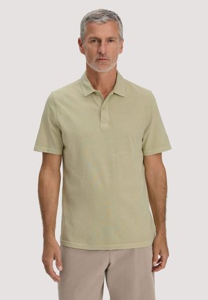 Middelbare man met kort grijs haar en baard, gekleed in een lichtgroene polo met korte mouwen en beige broek, staand tegen een effen achtergrond.