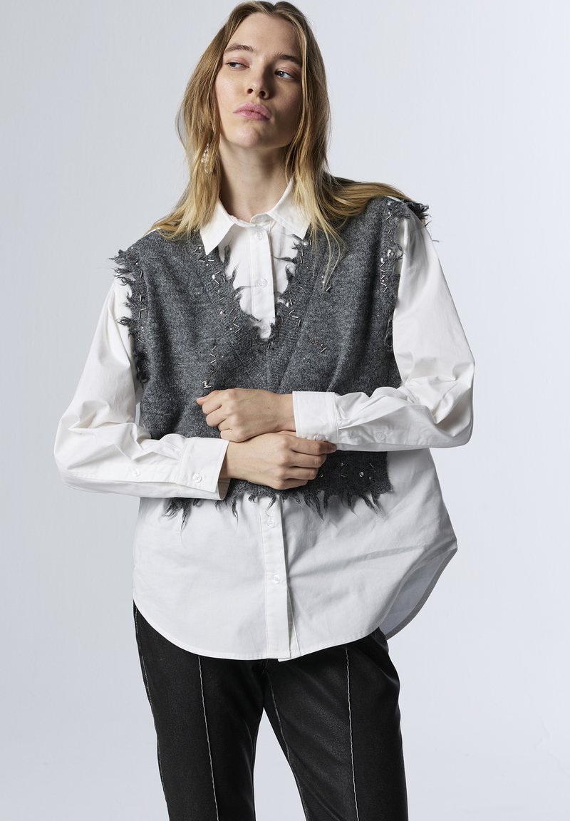 Gilet grigio con dettagli frangiati e hardware silver, sovrapposto a una camicia bianca con bottoni, abbinato a pantaloni neri con cuciture bianche.