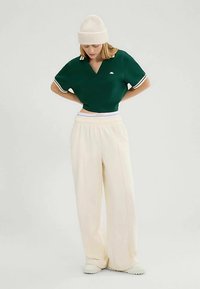 Groen cropped polo van katoen met witte bies; gecombineerd met crèmekleurige wijde broek. Model draagt een geribde beige muts voor warmte.