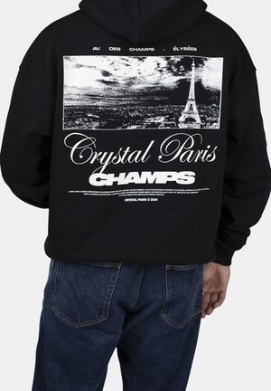 Persoon draagt een zwart hoodie met een witte afbeelding van de Eiffeltoren en de tekst "Crystal Paris Champs" op de achterkant, gecombineerd met een blauwe spijkerbroek.