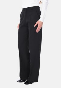 Pantaloni neri a gamba larga realizzati in tessuto morbido, con chiusura a bottone e passanti per cintura, abbinati a scarpe nere con punta.