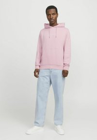 Pink hooded sweatshirt with drawstrings, front pocket, and white text logo, paired with light blue baggy jeans and white sneakers.