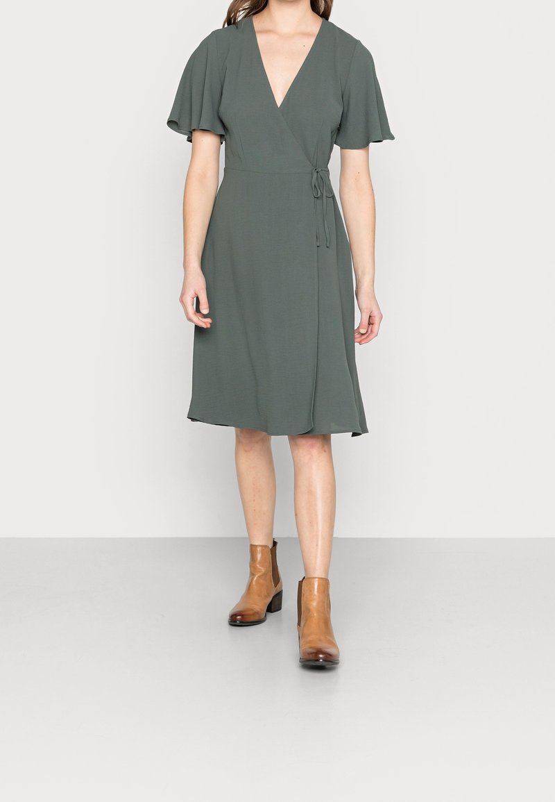 Tummanvihreä kietaisumekko, jossa on syvä V-kaula-aukko, lyhyet kelluvat hihat ja solmittava vyötärö. Mekossa on polveen ulottuva helma ja sileä kangas. Yhdistetty ruskeisiin nilkkasaappaisiin.