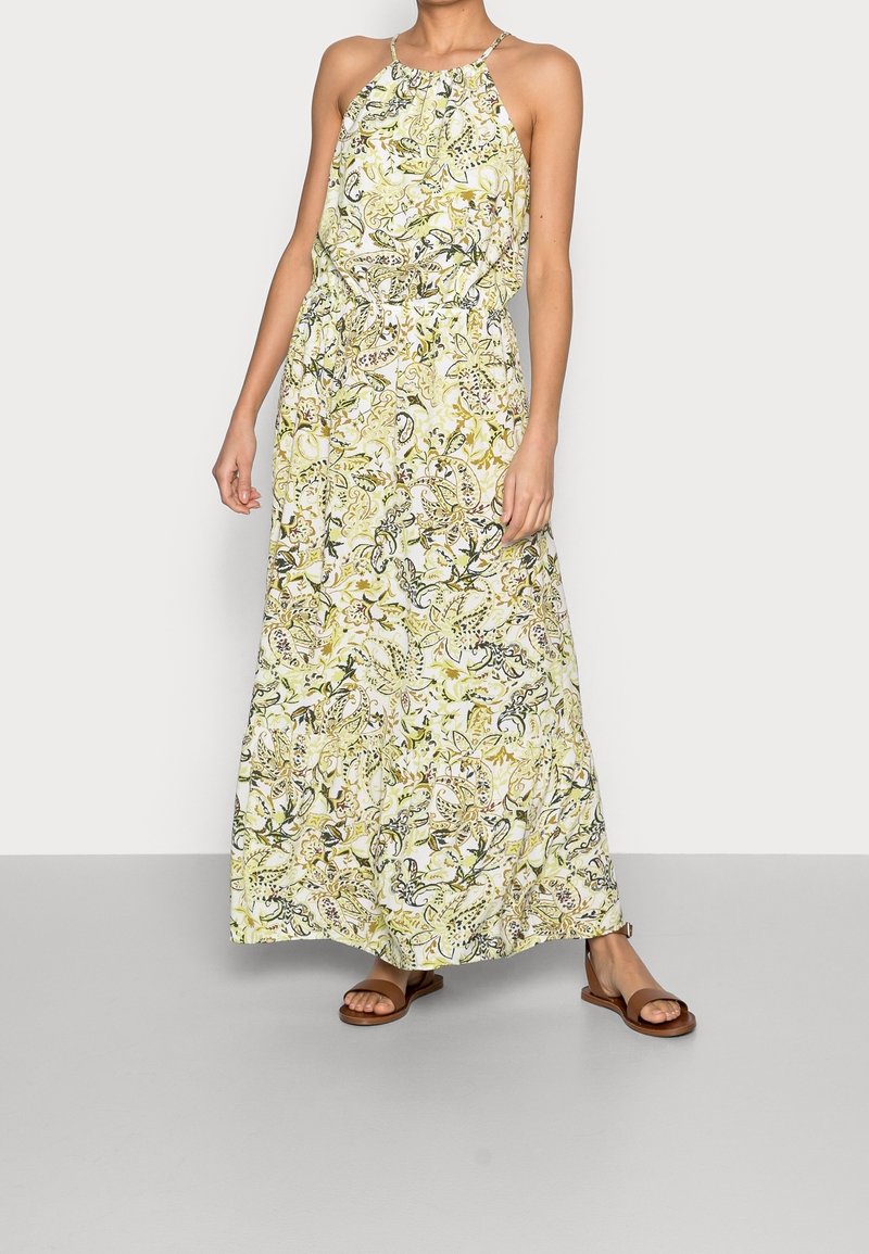 Femme portant une robe sans manches, col halter, longueur cheville, avec un motif floral paisley vert et beige et des sandales plates marron.